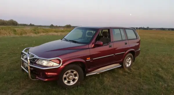 Ciemnoczerwony Daewoo Musso z orurowaniem z przodu stoi na trawiastym polu. W tle jezioro i księżyc.