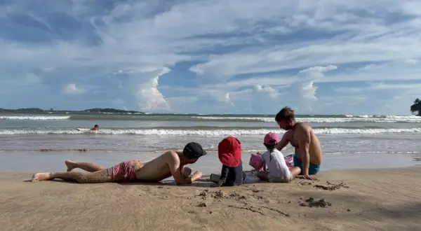 Familj njuter av en solig dag på stranden i Sri Lanka. En man ligger i sanden medan en annan leker med barnen vid havet.