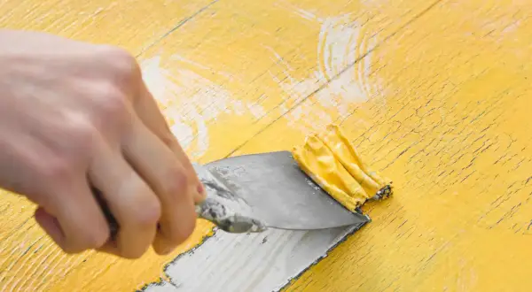 Mano raspando pintura amarilla vieja con una espátula. El decapado de pintura revela madera debajo.