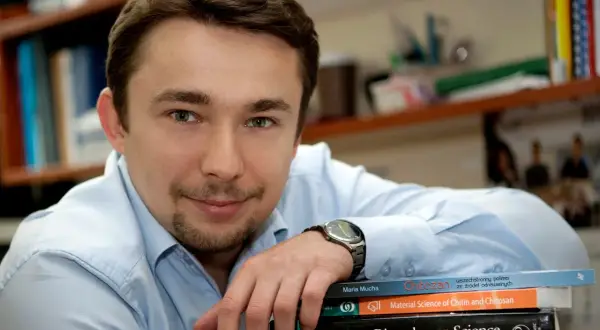 Mężczyzna w jasnej koszuli opiera rękę na stosie książek, w tym na tej zatytułowanej "Chitozan".