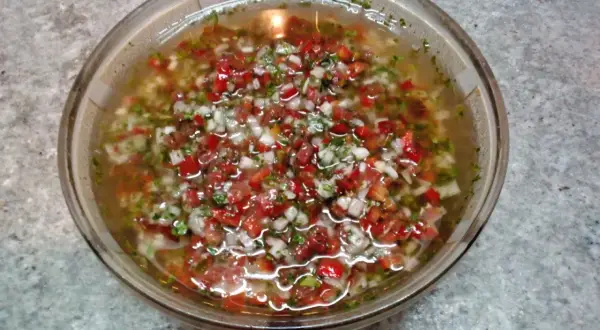 Un tazón de salsa criolla, con trozos picados de tomate, cebolla y pimiento rojo, flotando en un líquido claro.