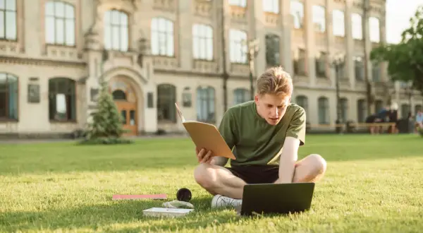 Joven estudiando en el césped frente a un edificio histórico, con un libro y portátil. Un momento de aprendizaje y **intercambio cultural** en un entorno inspirador.