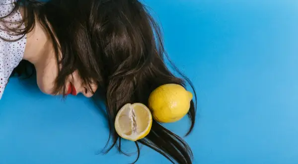En person med mörkt hår och röda läppar ligger på en blå bakgrund. Två citroner, en hel och en delad, ligger bland håret, som om de används för att bleka håret med citron.