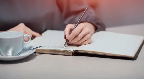 Dłoń pisze w notesie, szukając inspiracji, jak napisać intencję za zmarłego wzór. Obok filiżanka kawy.