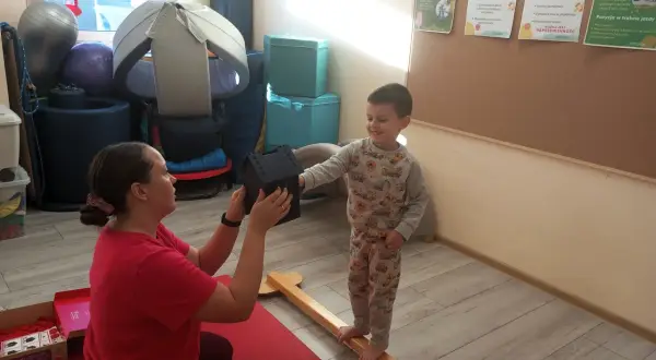 Fizjoterapeutka podaje chłopcu domek, który stoi na drewnianej belce.