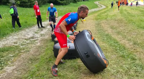 Uomo con treccia si allena al parco, spingendo un grande gonfiabile nero. Altri partecipanti corrono su un sentiero erboso.