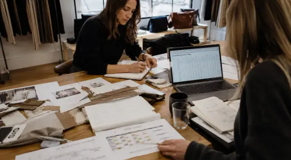 Dos mujeres revisan proyecciones de ventas y calendarios de producción. Una escribe en una libreta, la otra señala un documento. Se preguntan qué significa sin stock.