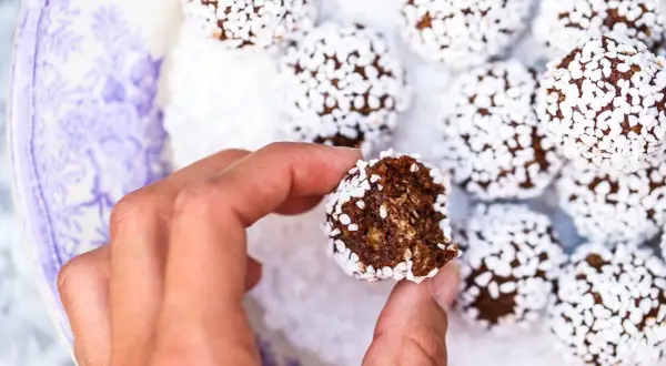 En hand håller en bit av nyttiga chokladbollar, rullade i kokosflingor, på en vacker lila-vit tallrik.