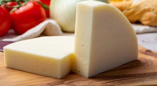 Zwei Stücke heller Käse auf einem Holzbrett, daneben Tomaten und Fenchel.