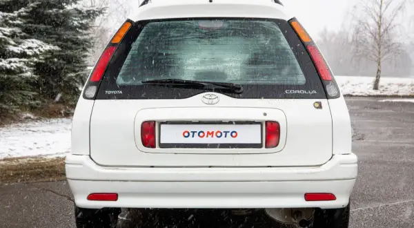 Biała Toyota Corolla E11 kombi z napisem "OTOMOTO" na tablicy rejestracyjnej, stoi na mokrym asfalcie podczas opadów śniegu.