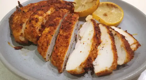 Skivad panerad kyckling med kryddig yta, serverad med grillade citronklyftor på en grå tallrik.