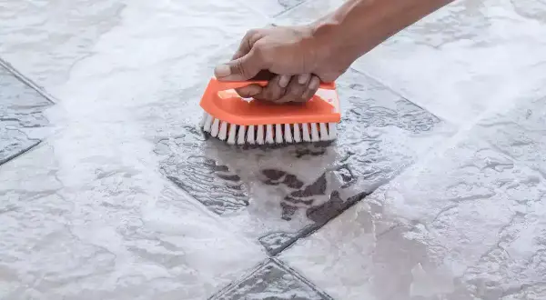 Main tenant nettoie une tache acide sur carrelage avec une brosse orange.