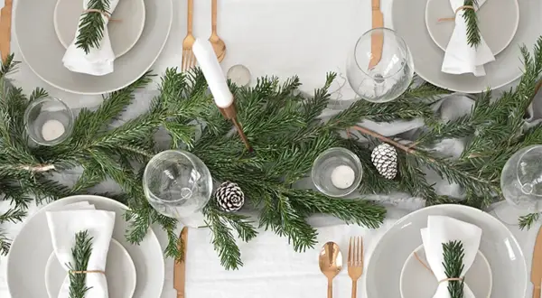 Sencillo decoracion mesa navidad con ramas de pino, piñas y velas. Vajilla gris, servilletas blancas con detalle de romero y cubertería dorada.
