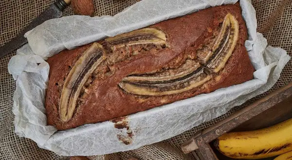 Nybakad banankaka i form, toppad med bananskivor. Valnötter och en gammal kniv ligger bredvid. Perfekt att baka med banan!