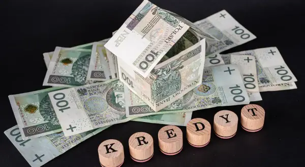Dom z banknotów 100 zł symbolizuje kredyt hipoteczny. Drewniane pieczątki układają się w napis "KREDYT", sugerując potrzebę restrukturyzacji.