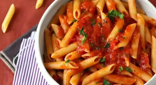 Penne all'arrabbiata w białej misce, polane pikantnym sosem pomidorowym z natką pietruszki. Obok leżą suche makarony.