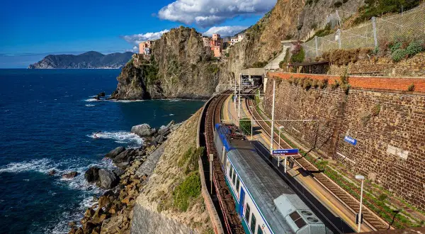 Ein Zug fährt entlang der Küste Italiens am Meer entlang. Bunte Häuser kleben an steilen Felsen.