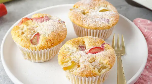 Drei saftige Rhabarber Muffins, mit Puderzucker bestäubt, auf einem weißen Teller. Ein goldener Gabel liegt daneben.
