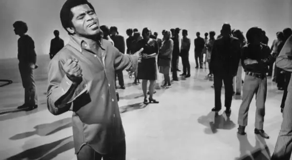 James Brown śpiewa na scenie, otoczony przez publiczność.