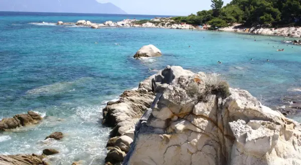 Słoneczne Chalkidiki plaże z turkusową wodą, skałami i ludźmi kąpiącymi się w morzu. W oddali widać góry.