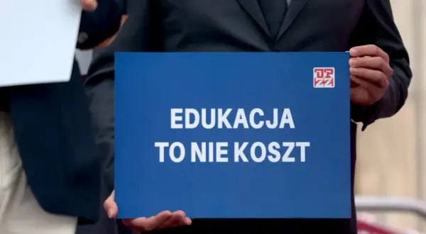 Osoba trzyma niebieską tabliczkę z napisem "EDUKACJA TO NIE KOSZT". Domaga się podwyżki dla nauczycieli 2026.
