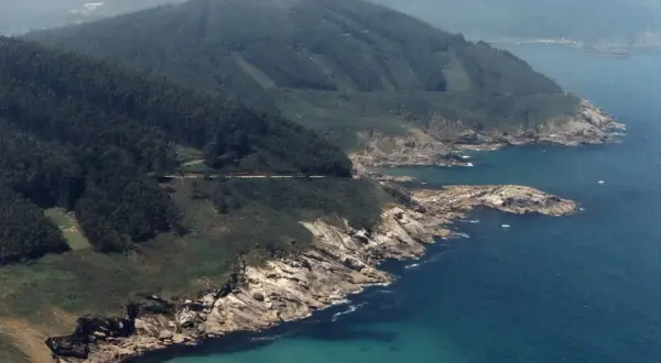 Costa rocosa y verde con el mar azul turquesa. Se ve una carretera serpenteando por la ladera. La playa Portonovo Viveiro es un paraíso natural.