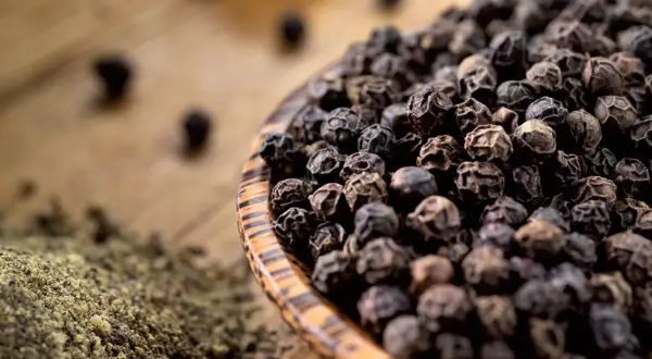 Schwarze Pfefferkörner in einer Holzschale, daneben gemahlener Pfeffer. So wird Pfeffer hergestellt: Ernte, Trocknung und Mahlung.