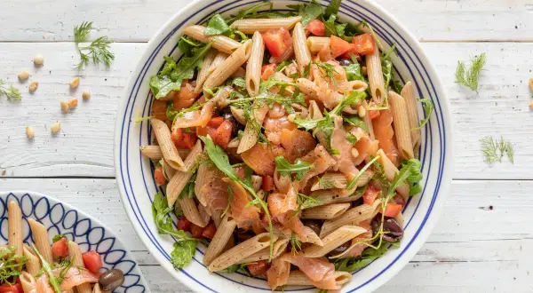 Pasta fredda con salmone affumicato, pomodorini, rucola e olive nere, guarnita con aneto fresco.
