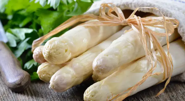 Un bouquet d'asperges blanches, prêtes à être poêlées, est noué avec une ficelle sur un tissu. Des herbes fraîches et un couteau sont visibles en arrière-plan.