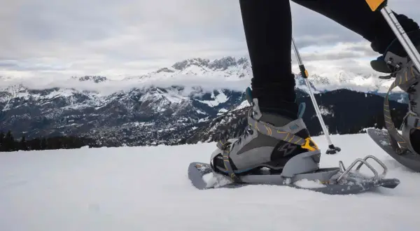 Una persona con ciaspole cammina sulla neve, con montagne innevate sullo sfondo.