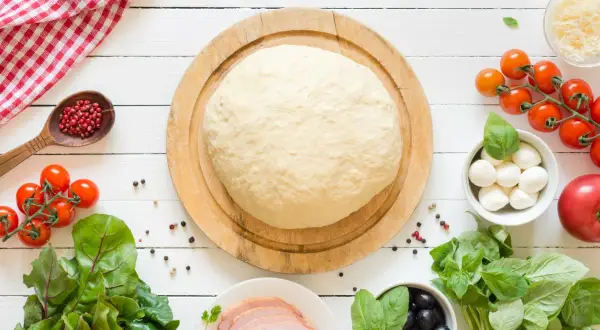 Surowe ciasto na pizzę z pomidorkami, mozzarellą, oliwkami i bazylią na białym tle.