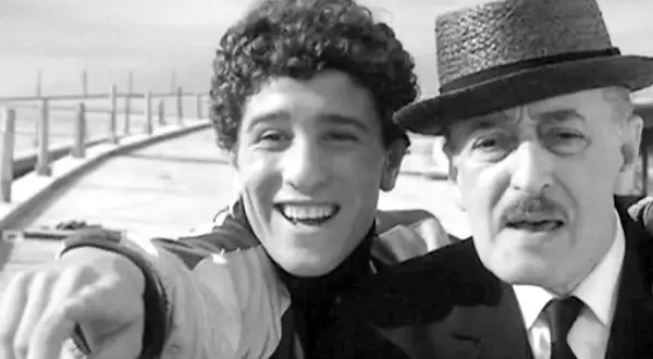 Un giovane sorridente con riccioli e un uomo anziano con cappello e baffi, un'immagine che trasmette un senso di complicità e un significato di legame generazionale.
