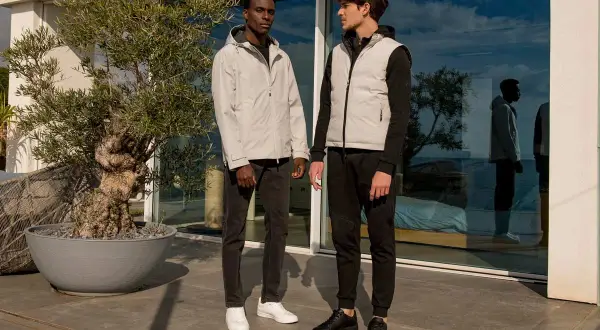 Due uomini eleganti, uno con giacca grigia e jeans, l'altro con gilet grigio e pantaloni sportivi neri, posano all'esterno.