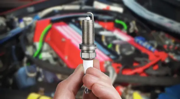Une bougie d'allumage tenue en main, possible cause d'une voiture qui broute au démarrage. Le moteur est flou en arrière-plan.