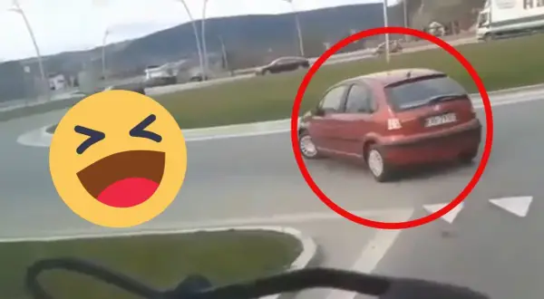 Czerwony samochód próbuje zawrócić na rondzie, ale robi to na lewo, co wygląda na niezbyt przemyślane. Obok śmieje się emotikon.