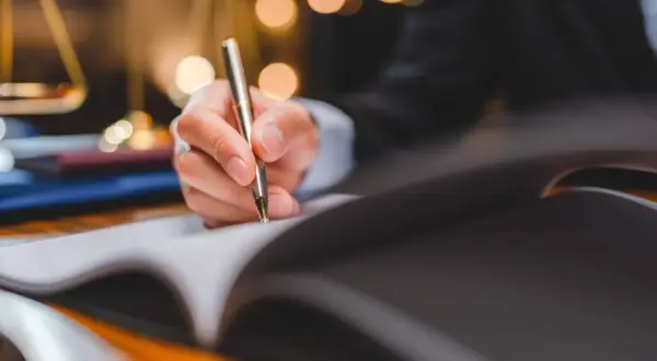 Dłoń pisze piórem w otwartym notatniku. W tle widać wagę sprawiedliwości i rozmyte światła, sugerujące formalne dokumenty, jak promesa zatrudnienia wzór.