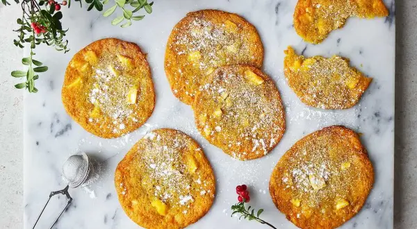 Sju gyllene saffranscookies med vit choklad, pudrade med florsocker, ligger på ett marmorfat. En bit är tagen från en cookie.