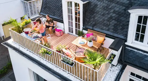 Ein Paar genießt ein Grillfest auf ihrem neu gestalteten Balkon. Der Balkon ist mit Holzmöbeln, Pflanzen und Dekorationen geschmückt.