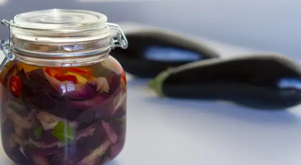 Berenjenas en escabeche, cortadas y marinadas en un frasco de vidrio, con berenjenas frescas desenfocadas al fondo.
