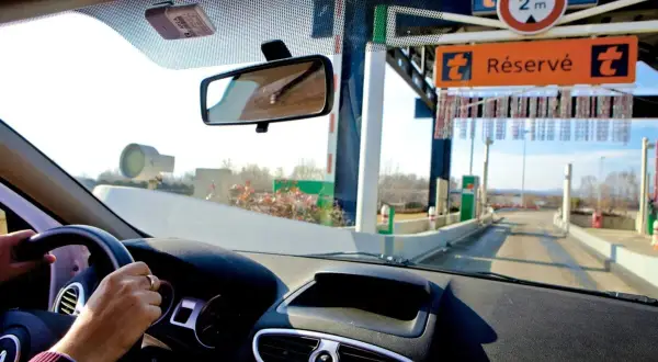Vue depuis l'intérieur d'une voiture approchant une voie de péage. Le tableau de bord et le volant sont visibles. Un badge de télépéage est fixé au pare-brise. Le panneau "Réservé" indique comment ça fonctionne.