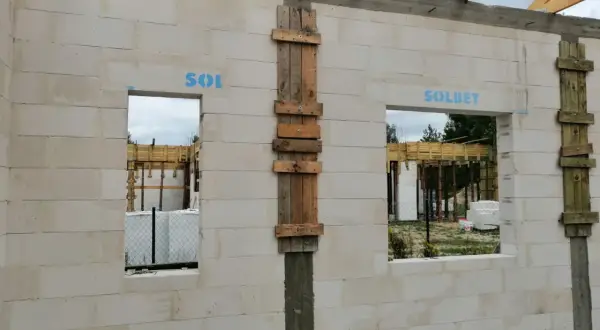 Budowa domu z bloczków betonowych, widoczne okna i drzwi z tymczasowym nadprożem, drewniane elementy konstrukcyjne.