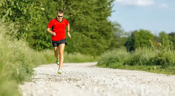 Mann in rotem Shirt und schwarzen Shorts absolviert einen Fartlek Lauf auf einem Schotterweg, umgeben von grüner Natur.
