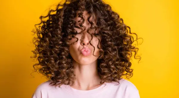 Mujer con un espectacular pelo rizado fino y castaño, haciendo un beso contra un fondo amarillo vibrante.