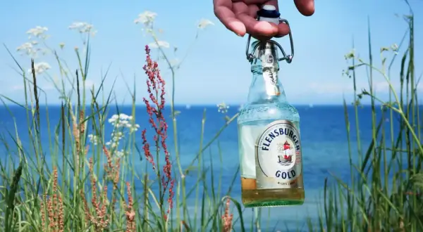 Eine Flensburger Gold Flasche wird vor der Küste gehalten. Der leichte Bier Geschmack passt perfekt zum Sommer.