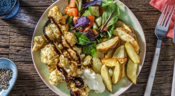En tallrik med rostad blomkål i ugn, klyftpotatis och en fräsch sallad. Blomkålen är täckt med ströbröd och ringlad med en mörk sås.