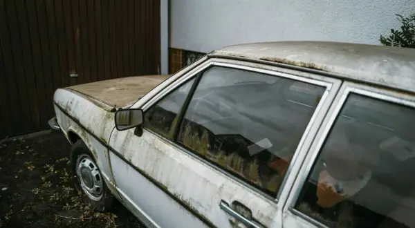 Vieille voiture blanche, couverte de poussière et de mousse, devant un garage. Un symbole de l'interdiction voiture thermique.