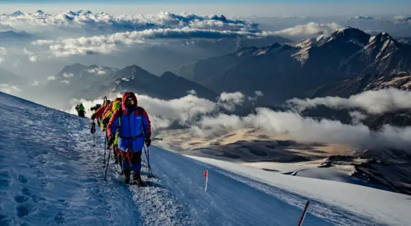 Grupa alpinistów wspina się po ośnieżonym zboczu, podziwiając majestatyczne góry z Elbrusem w tle. Chmury unoszą się poniżej, tworząc baśniowy krajobraz.