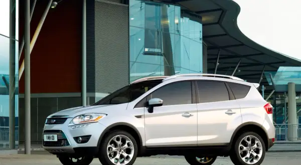 Biały Ford Kuga 2010 zaparkowany przed nowoczesnym budynkiem z przeszkloną fasadą.