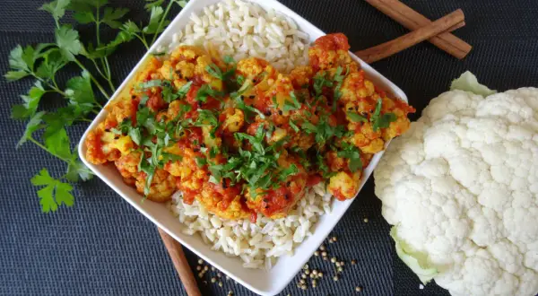 Aksamitne curry z kalafiora podane z ryżem, udekorowane świeżą natką pietruszki i czarnuszką. Obok leży świeży kalafior i laski cynamonu.