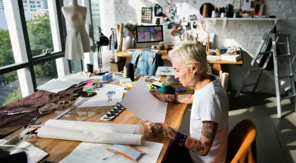 A designer sketches at a table filled with fabric and patterns, a scene that captures the essence of what is an artist residency: a dedicated space for creative exploration and development.
